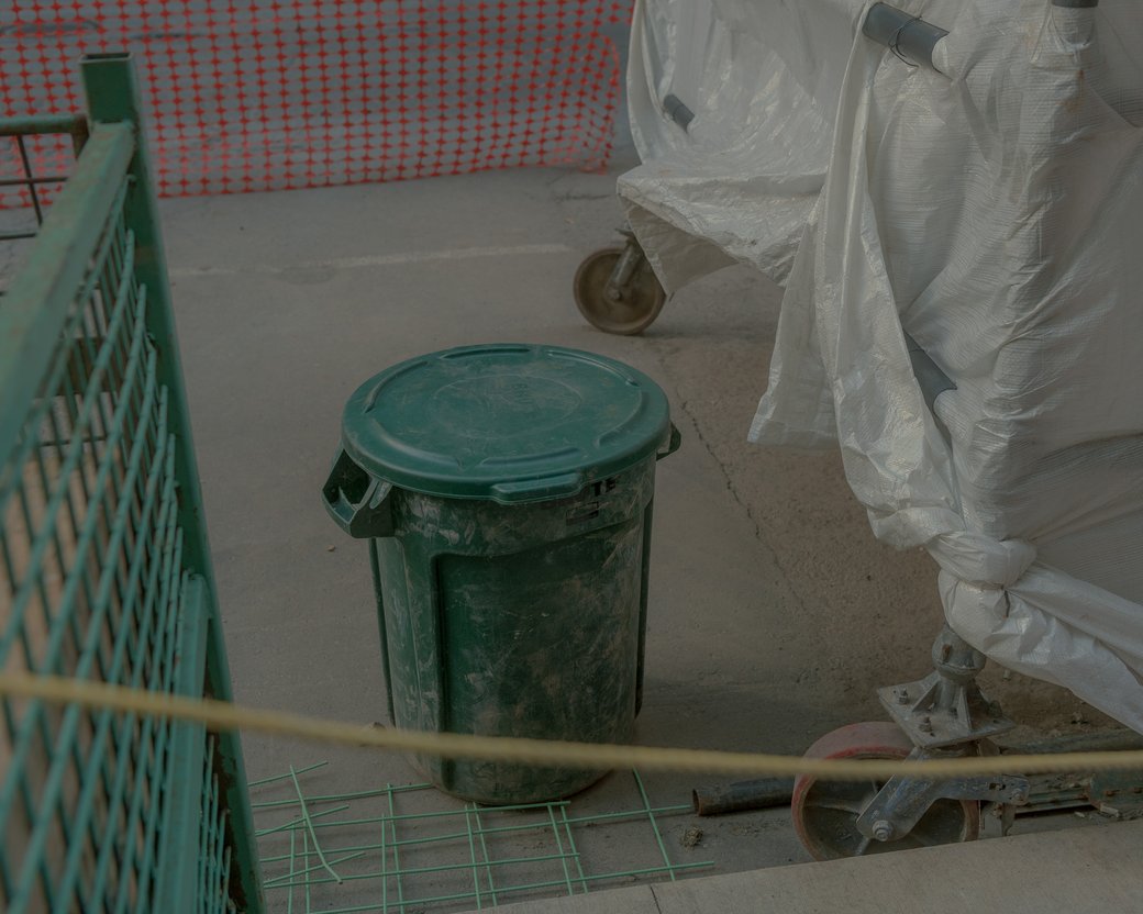 Green trash bin on a concrete construction site beside orange safety netting.