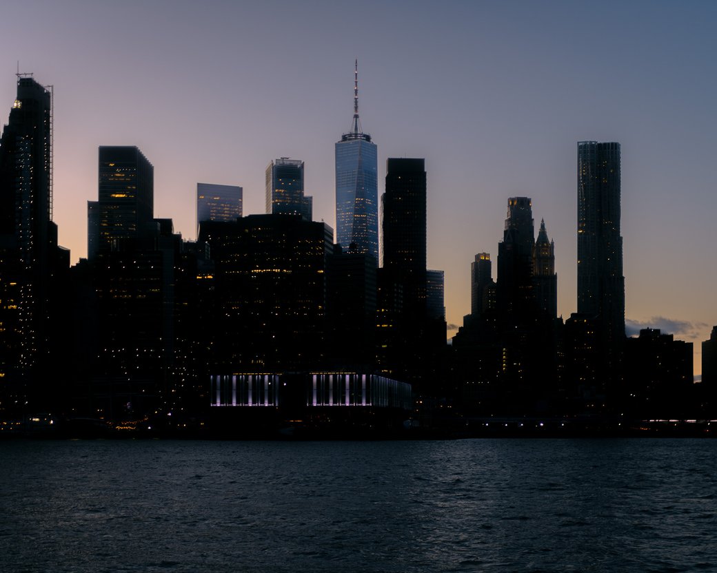 City skyline at dusk with One World Trade Center rising above dark buildings across the water.