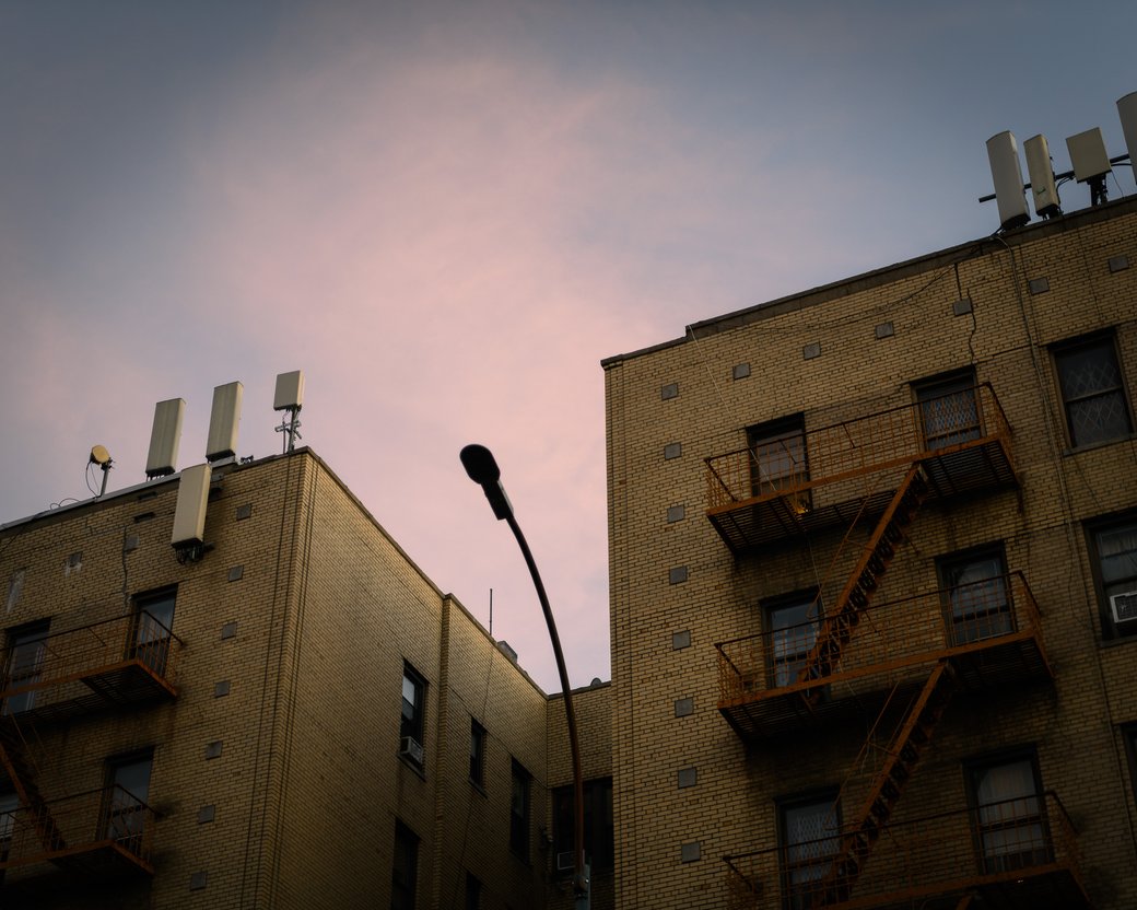 Brick apartment buildings with fire escapes and rooftop antennas against a pink evening sky.