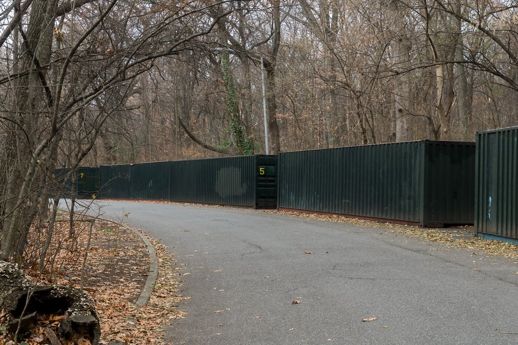 Curving paved path through leafless woods beside a long dark green metal shipping container.